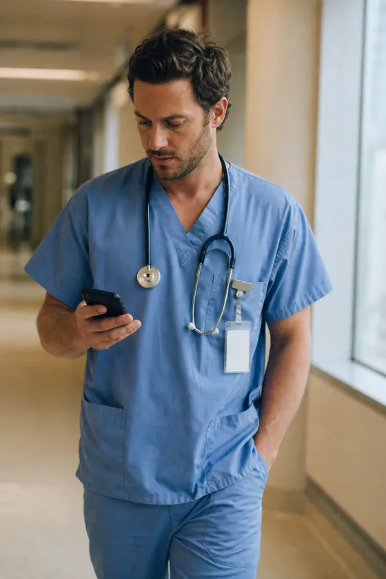 A doctor in light blue scrubs checking his phone in a hospital corridor